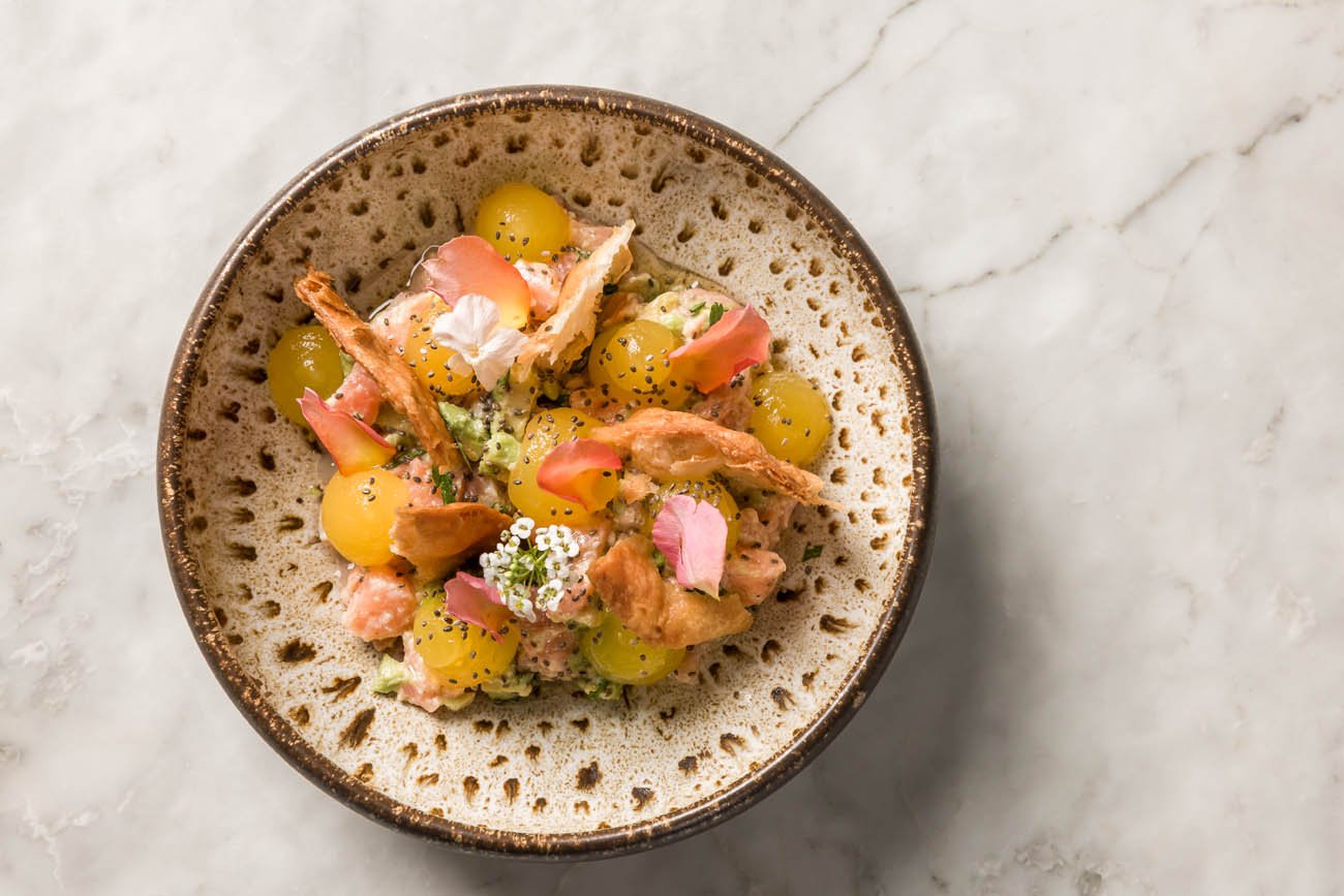 Prato de salada com uvas verdes, flores comestíveis, pétalas de rosa e pedaços de frango em molho, servido em tigela de cerâmica