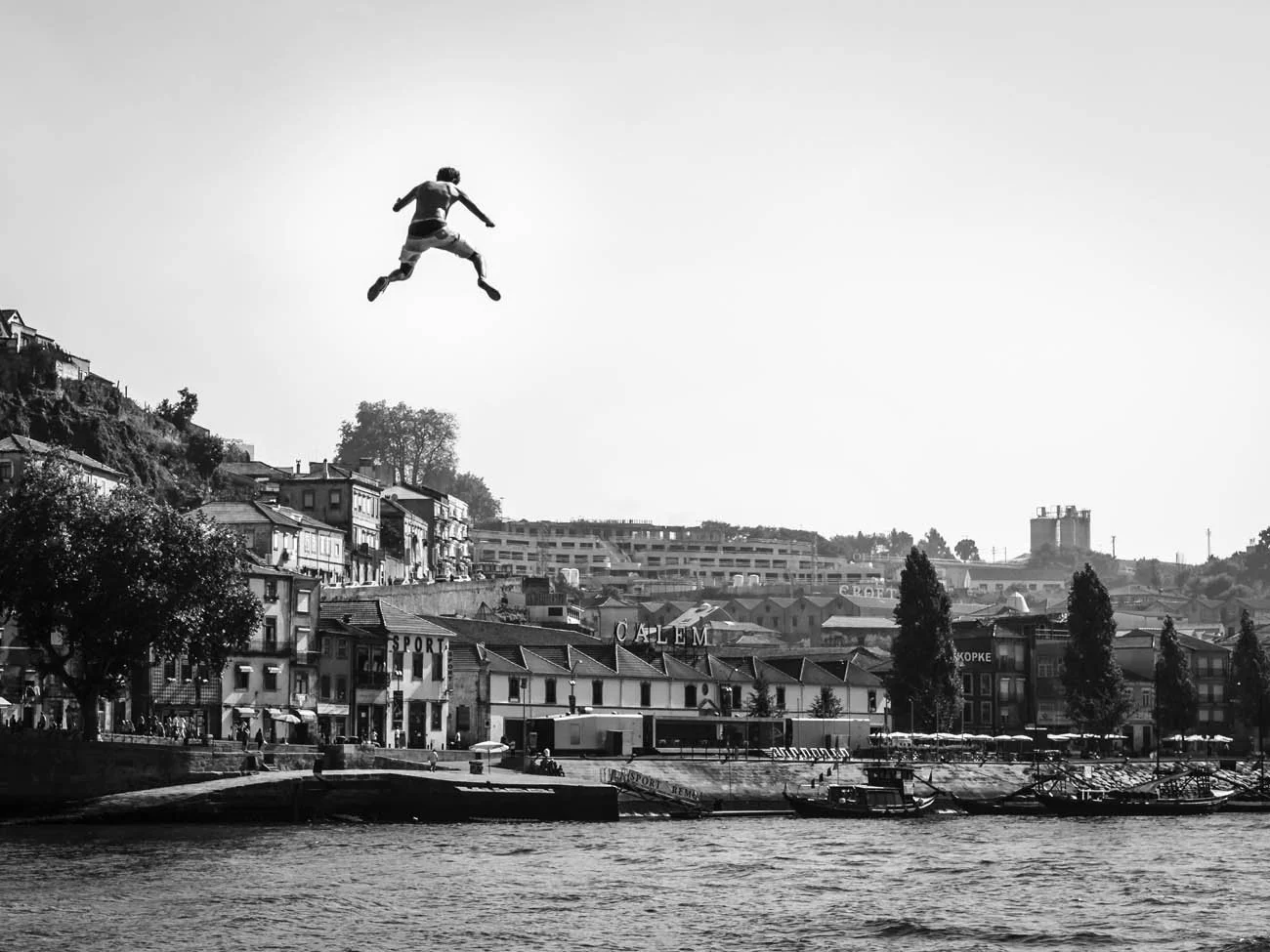 Pessoa pulando de um local elevado em direção a uma área com casas, árvores e barcos no rio, em uma paisagem urbana ao fundo, em preto e branco.