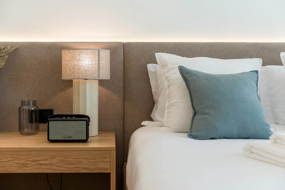 Cama com cabeceira acolchoada, travesseiro azul, travesseiros brancos, luminária de mesa, vaso de vidro marrom, rádio portátil, roupa de cama brancas na cama.