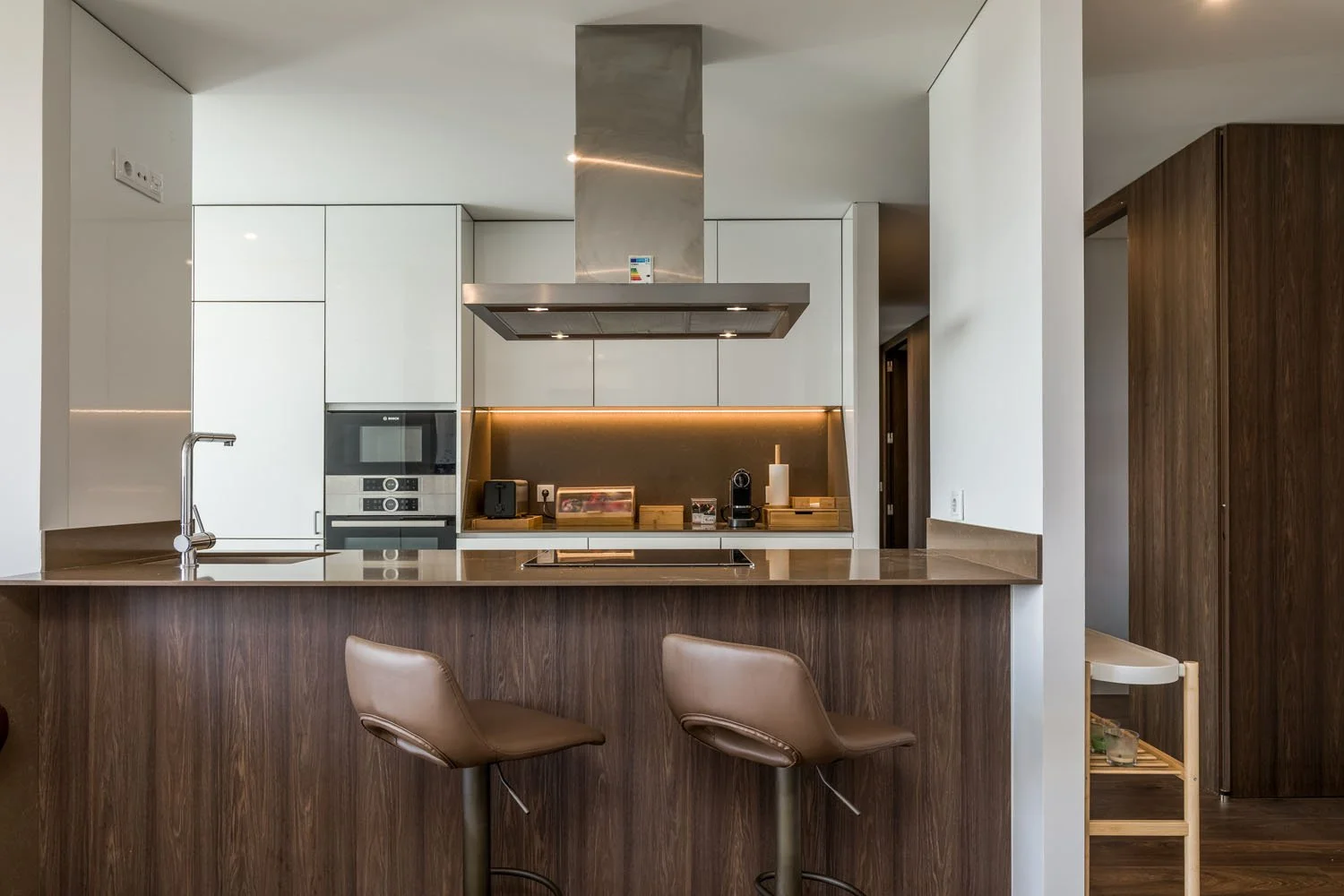 Cozinha moderna com ilha de madeira, bancos de couro, armários brancos, paredes com iluminação embutida, eletrodomésticos de aço inox e detalhes de madeira.