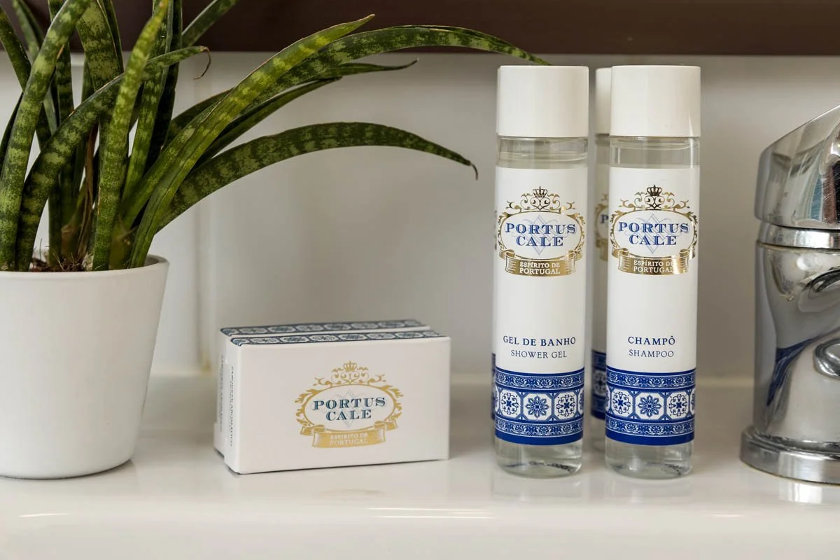 Itens de higiene pessoal incluindo sabonete, sabonete em pó, gel de banho e shampoo, ao lado de uma planta em um vaso branco, numa bancada de banheiro