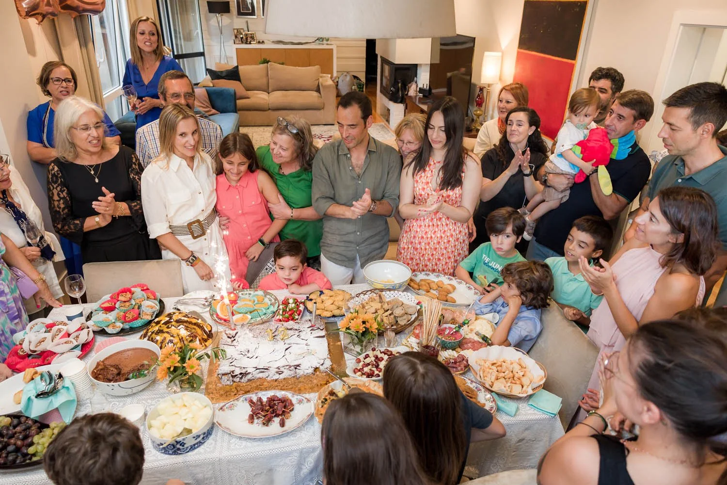 Reunião de aniversário com várias pessoas ao redor de uma mesa com bolos, doces e salgados, com uma criança no centro e muitos adultos e crianças acompanhando a celebração na sala de estar.