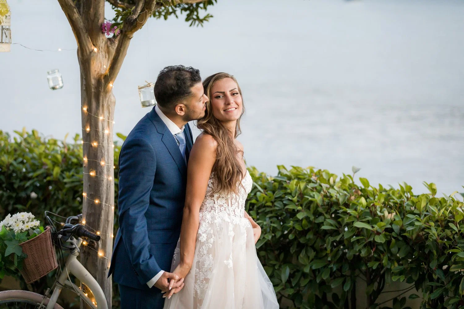 Um casal de noivos na praia, segurando as mãos, com o homem beijando a testa da mulher. Ela está sorrindo, usando vestido de casamento branco. Há um poste de madeira com luzes e uma bicicleta com cesta de plantas ao lado, com fundo de mar e vegetação