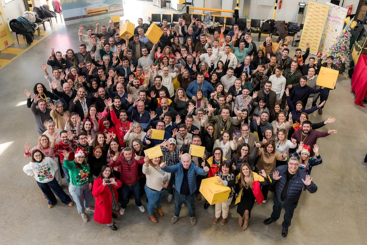 Grupo de várias pessoas sorrindo e acenando para a câmera em um ambiente interno, alguns segurando caixas e usando camisetas e roupas de comemoração.