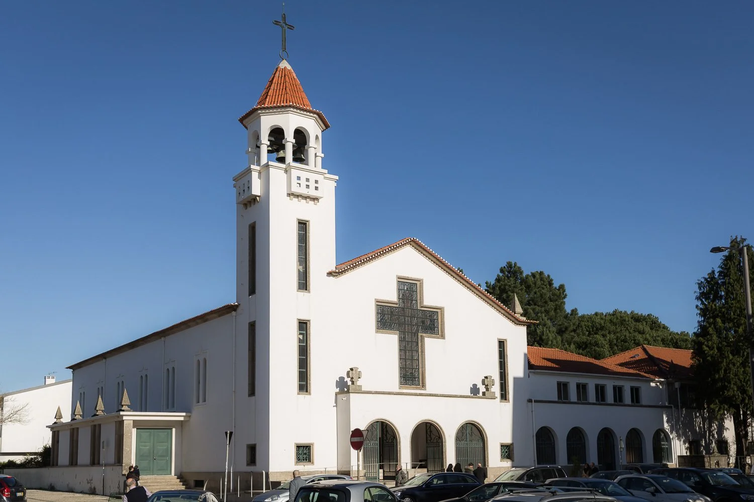Igreja branca com torre e cruz no topo, janelas altas e arco na entrada, veículos estacionados na frente e pessoas. Árvores ao fundo e céu claro.