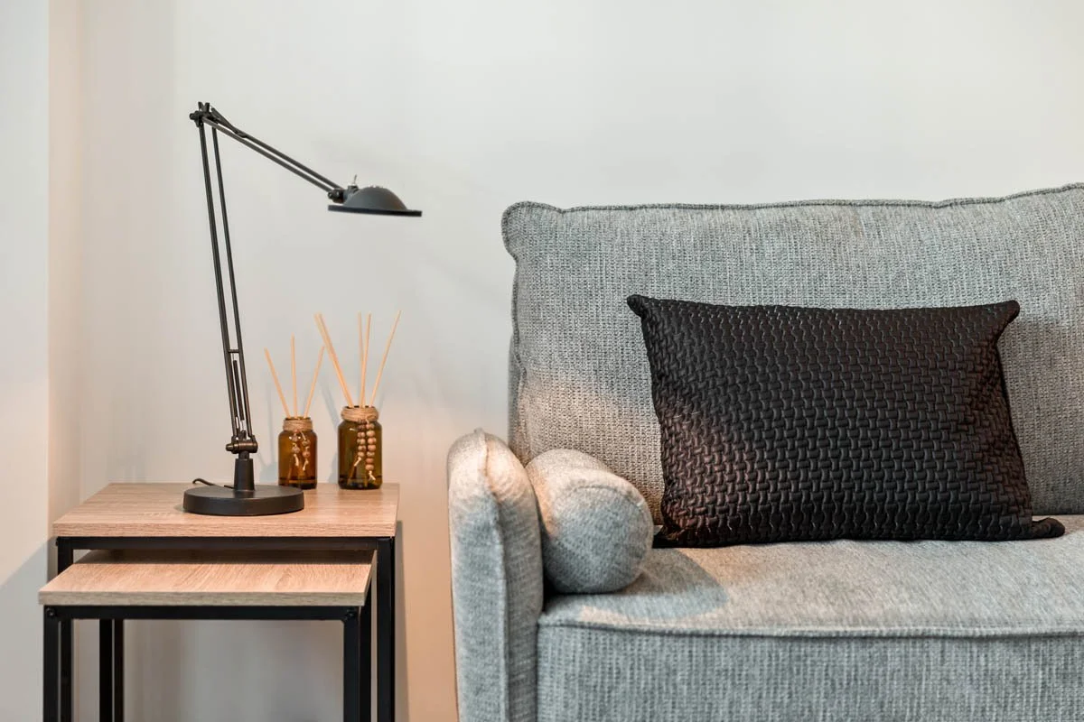 Sala de estar com sofá cinza, almofada preta e luminária de mesa preta ao lado de duas velas com frascos de vidro e varetas de difusor.