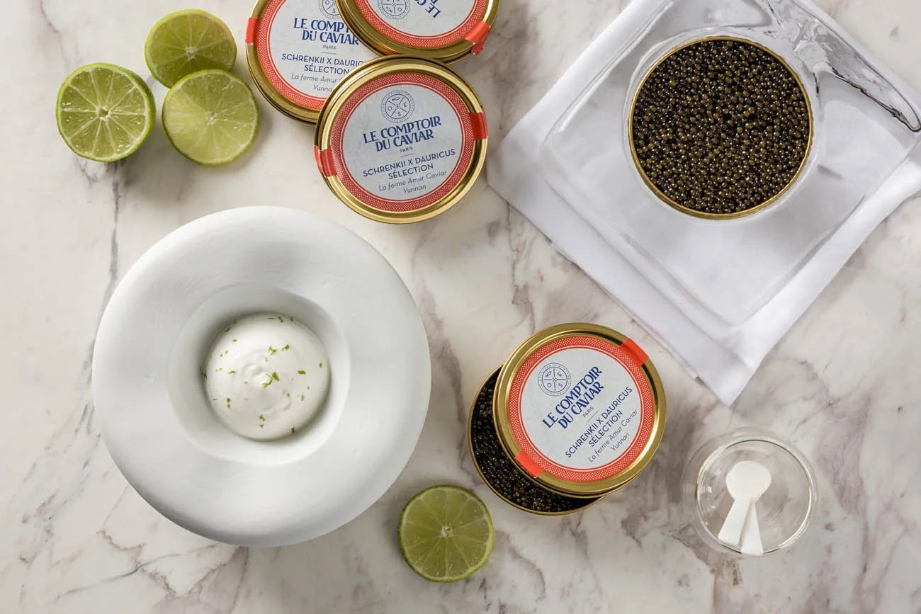 Latas de ovas de peixe, limões cortados ao meio, uma tigela com creme com raspas de limão, uma colher de metal, uma peneira com ovas de peixe e um recipiente de vidro pequeno com uma colher