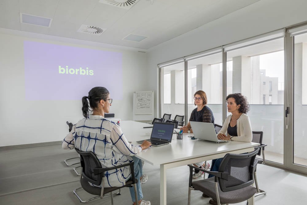 Reunião de três mulheres em uma sala de conferências com laptops, uma delas apresenta um slide que diz 'biorbis'.