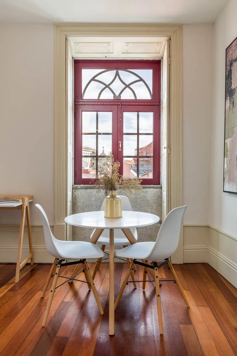 Sala de jantar com janela grande vermelha, mesa branca, três cadeiras brancas ao redor, vaso com flores secas no centro e piso de madeira