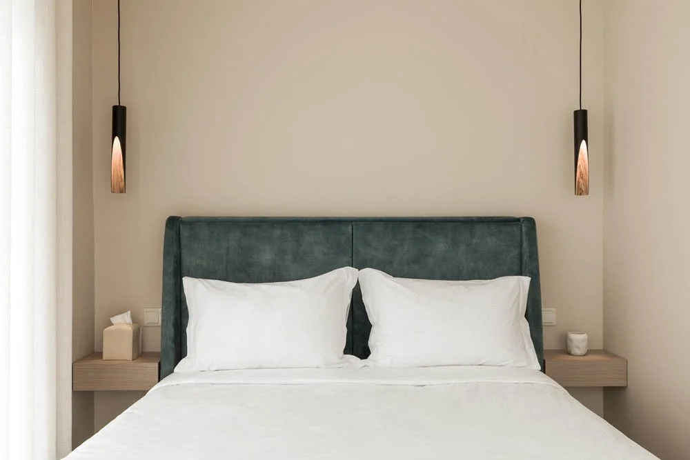 Cama de casal com cabeceira estofada verde em um quarto de estilo minimalista, com duas luminárias penduradas em ambos os lados da cama e uma parede de cor clara ao fundo.