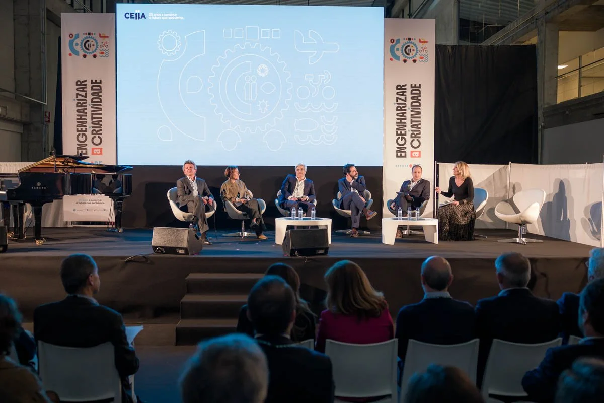 Painel de fundo com marcação do evento "Encengelharizar a Criatividade" pelo CEIIA. Pessoas participando de uma mesa redonda em um palco, com público assistindo.