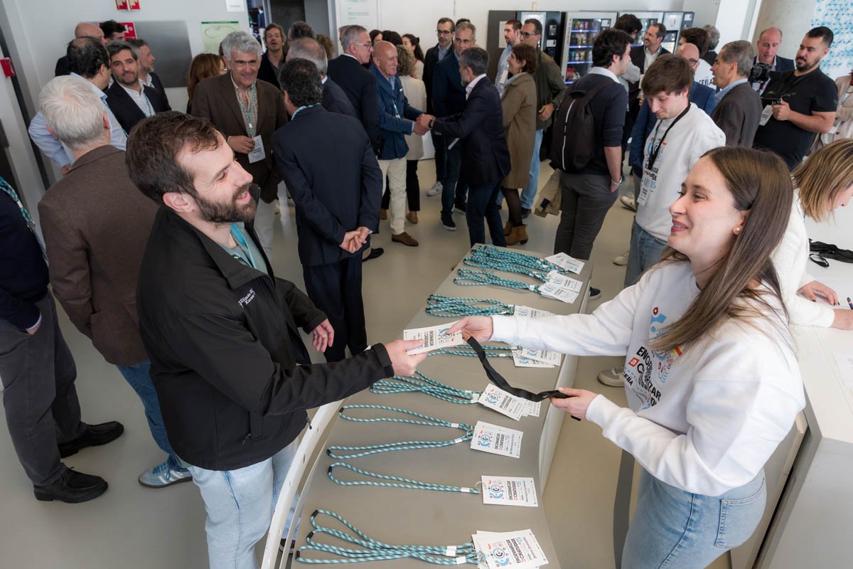 Pessoa entregando crachá para participante em evento de negócios, com várias pessoas conversando ao fundo.