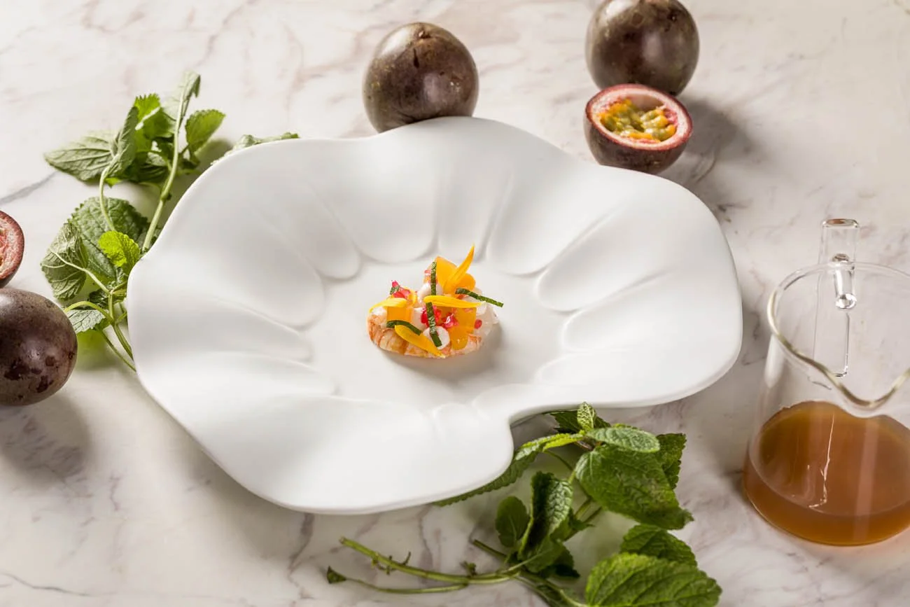 Prato pequeno de comida com decoração colorida, servido em uma tigela branca com formato de folha, ao redor há maracujás, folhas de hortelã e um recipiente com molho ou vinagre, tudo disposto sobre uma superfície de pedra clara.