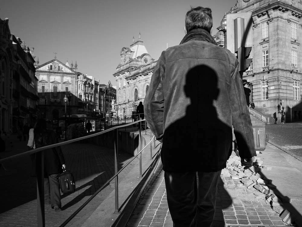 Homem com jaqueta escura caminhando na rua com sombra projetada na calçada, ao lado de edifícios históricos em uma cidade.