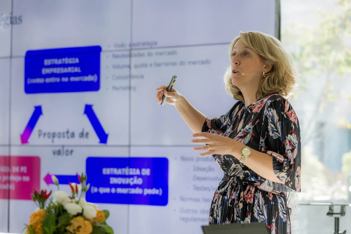 Mulher dando uma palestra ou aula, apontando para uma tela com um diagrama e texto relacionados a estratégias de marketing, com flores na frente.