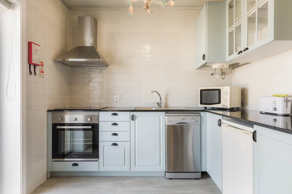 Cozinha com fogão elétrico, forno, micro-ondas, geladeira, armários brancos e uma pia com torneira, decorada com luminária de teto.