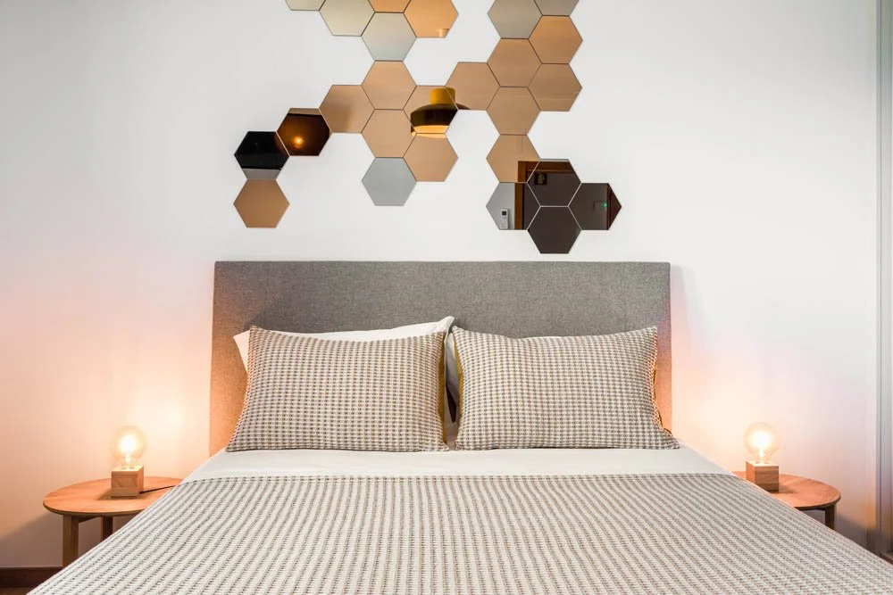 Quarto com cabeceira cinza, duas almofadas de roupa xadrez, duas lâmpadas de mesa acesas, parede com espelhos hexagonais em tons de bege e preto.
