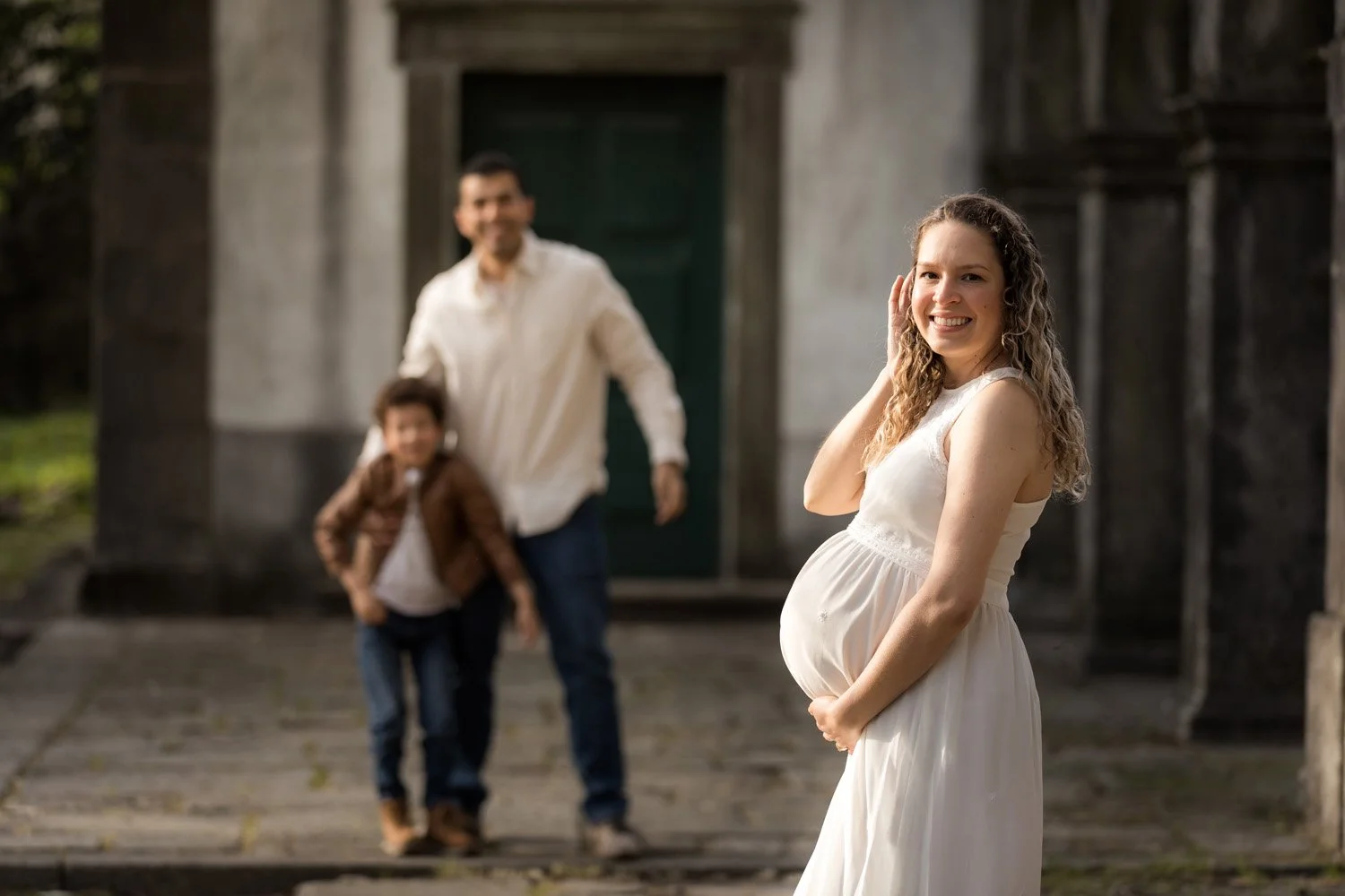 Mulher grávida sorridente posando ao ar livre com família ao fundo, incluindo um homem e uma criança, em uma cena de ensaio fotográfico.