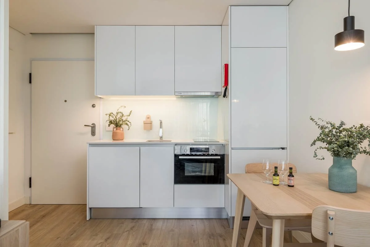 Cozinha moderna com armários brancos, forno embutido, pia com torneira, mesa de jantar de madeira com duas cadeiras, vaso grande com planta verde, bandeja com duas garrafas e taças de vinho, iluminação suspensa preta.