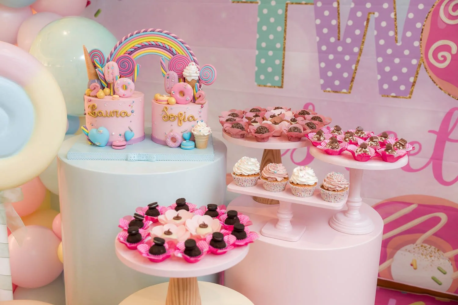 Mesa de celebração com diversos doces e bolos decorados para uma festa infantil. Dois bolos de aniversário rosa com decoração de arco-íris, nuvens, e nomes "Laura" e "Sofia". Pratos com cupcakes, brigadeiros e docinhos decorados com cores rosa, branc