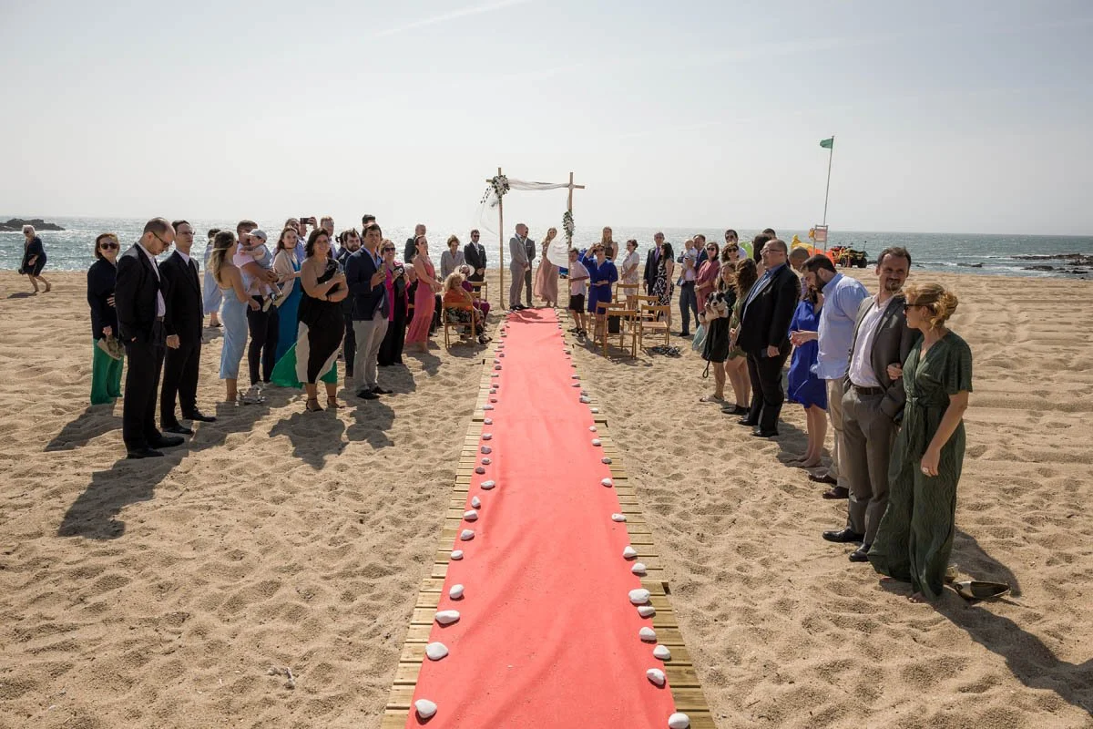 Casamento na praia com convidados, altar, tábua vermelha ao longo do caminho, pedras brancas decorativas, mar ao fundo.