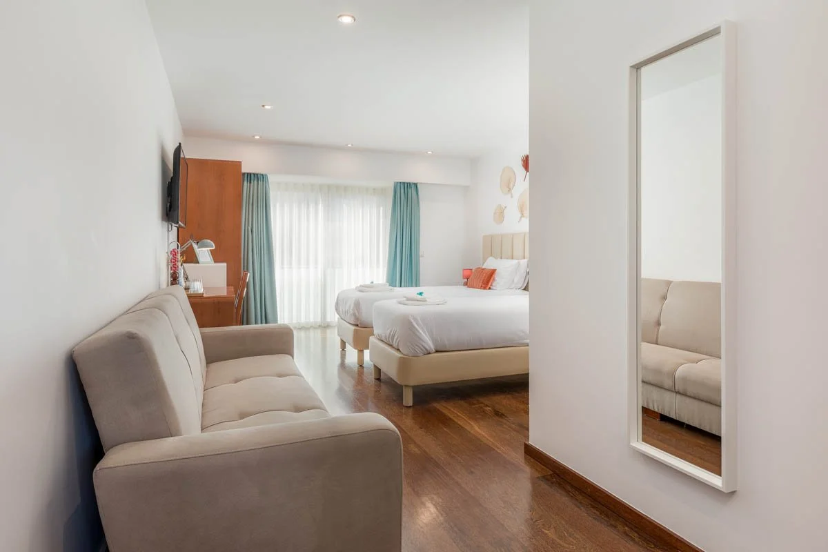 Quarto de hotel com duas camas de casal com roupas de cama brancas, sofá bege, espelho na parede, cortinas azuis e pastéis, televisão na parede e piso de madeira.