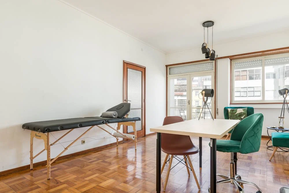 Sala de estar com móveis modernos, incluindo uma mesa, cadeiras, uma cama de massagem e luminárias de piso, com janelas grandes e piso de madeira.