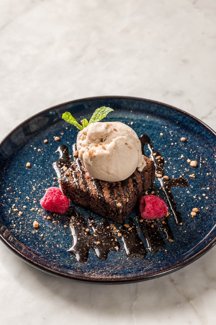 Sobremesa com bolo de chocolate, uma bola de sorvete, calda de chocolate, damascos, framboesas, e hortelã, em um prato azul escuro
