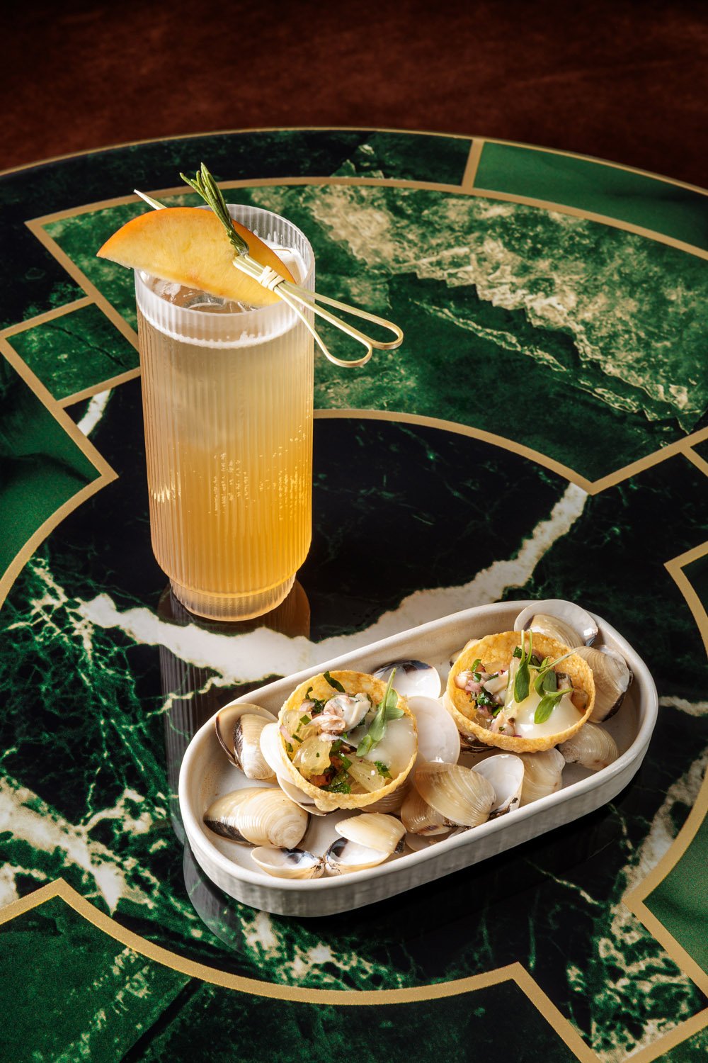 Copo de drink com fatia de fruta e ramos de alecrim, acompanhados de uma travessa com croquetes recheados e mariscos.