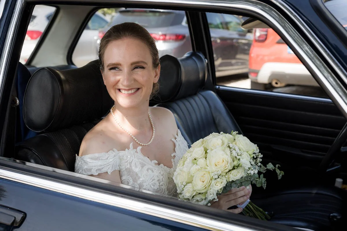 No interior de um carro, uma mulher sorridente usando vestido de noiva e segurando um buquê de flores brancas, com um fundo de outros carros estacionados.