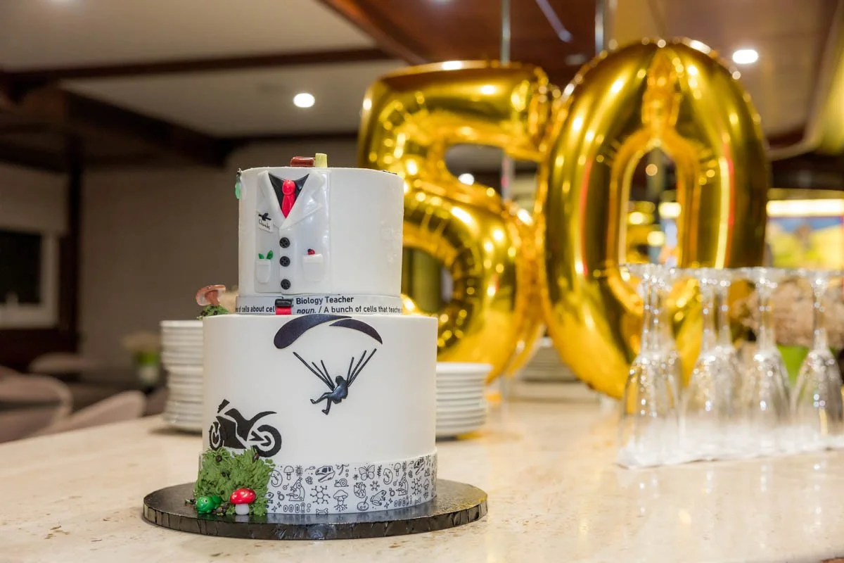 Bolo de casamento com decoração temática de biologia, incluindo um cientista de gravata e jaleco, uma pessoa pulando de paraquedas, uma moto e um paraquedista. Ao fundo, números grandes de balões dourados formando '50' e copos de vidro empilhados. Ce