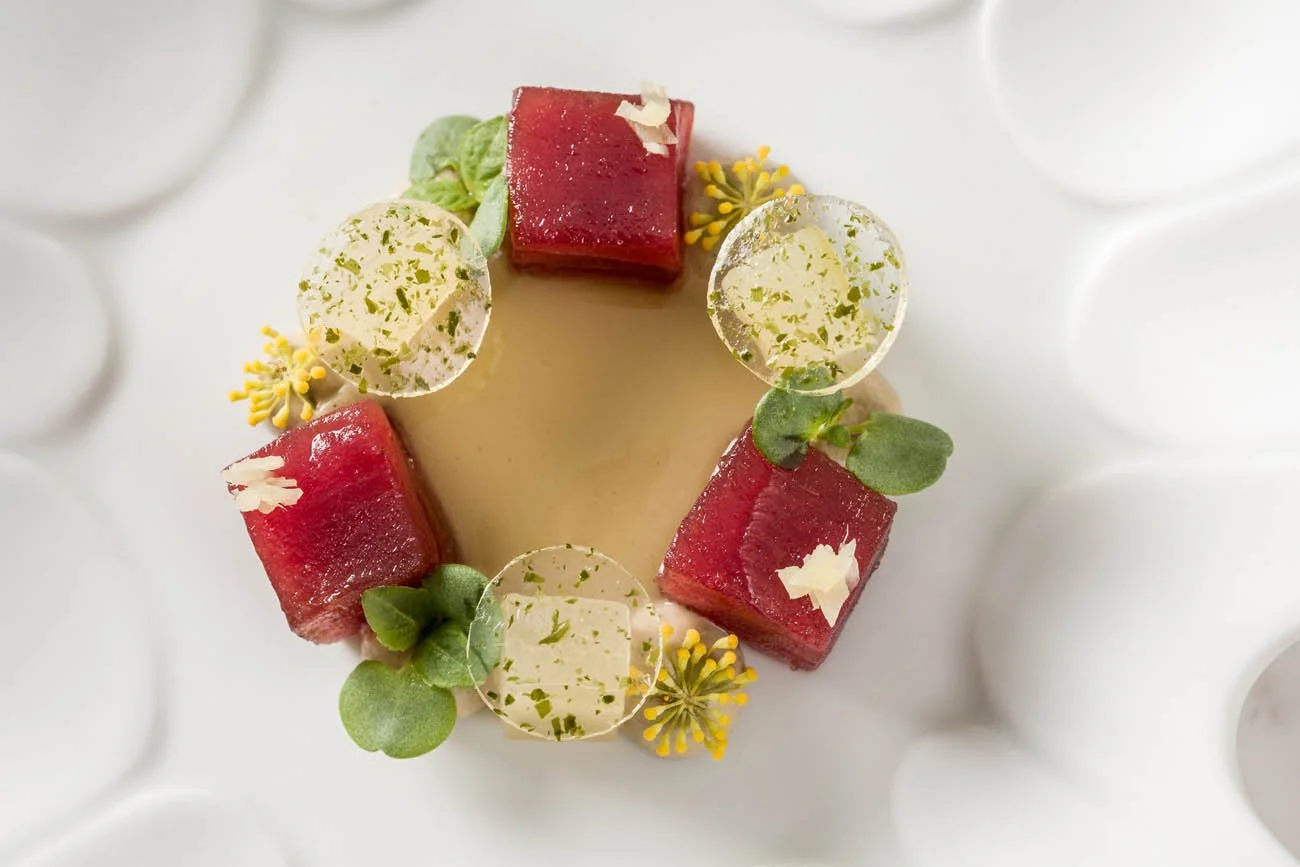 Sobremesa com cubos de gelatina vermelha, folhas verdes, pequenas flores amarelas e discos transparentes decorativos, disposta sobre um prato branco com relevo.