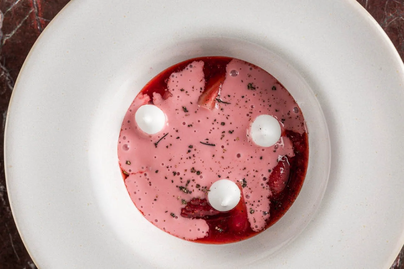 Sobremesa de morangos com creme de cor rosa, decorada com três bolas brancas, em um prato branco.