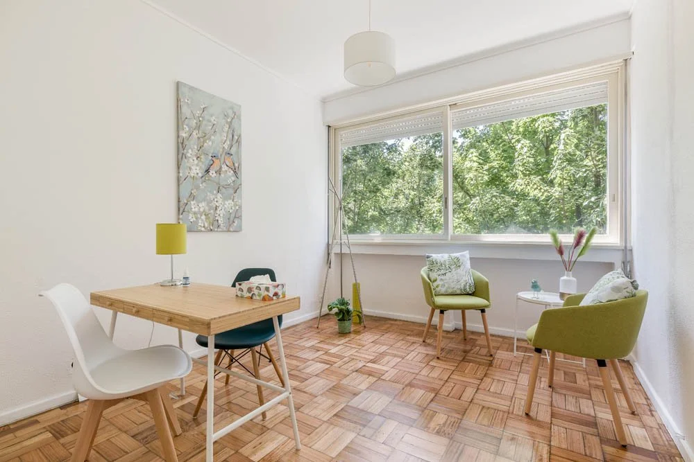 Sala de estar com sofá, duas poltronas, mesa de canto com vaso de plantas, janela grande com vista para árvores verdes, quadro na parede e piso de madeira.