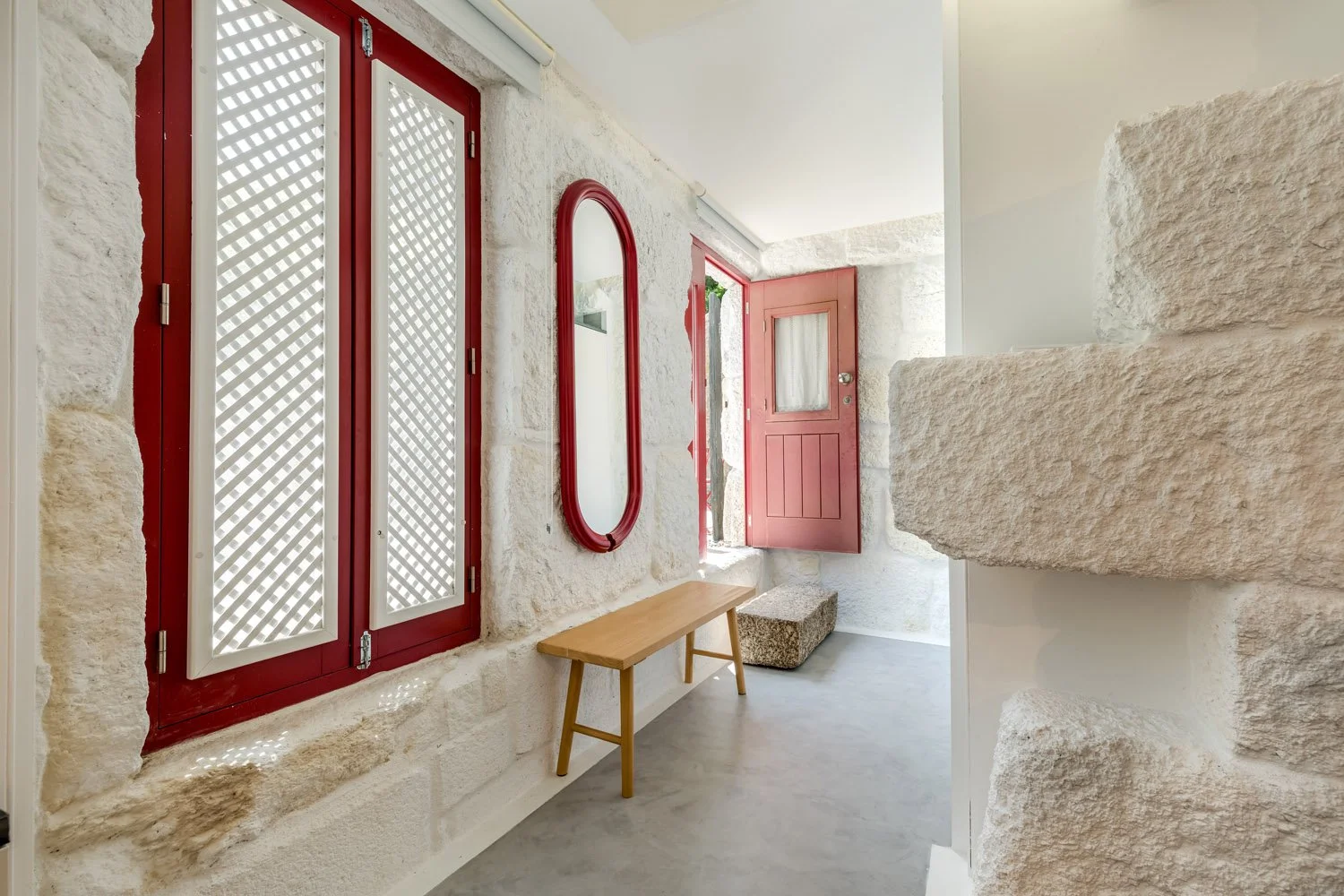 Entrada com paredes de pedra, janela e porta pintadas de vermelho, espelho de moldura vermelha, banco de madeira, piso de concreto e escada de pedra.