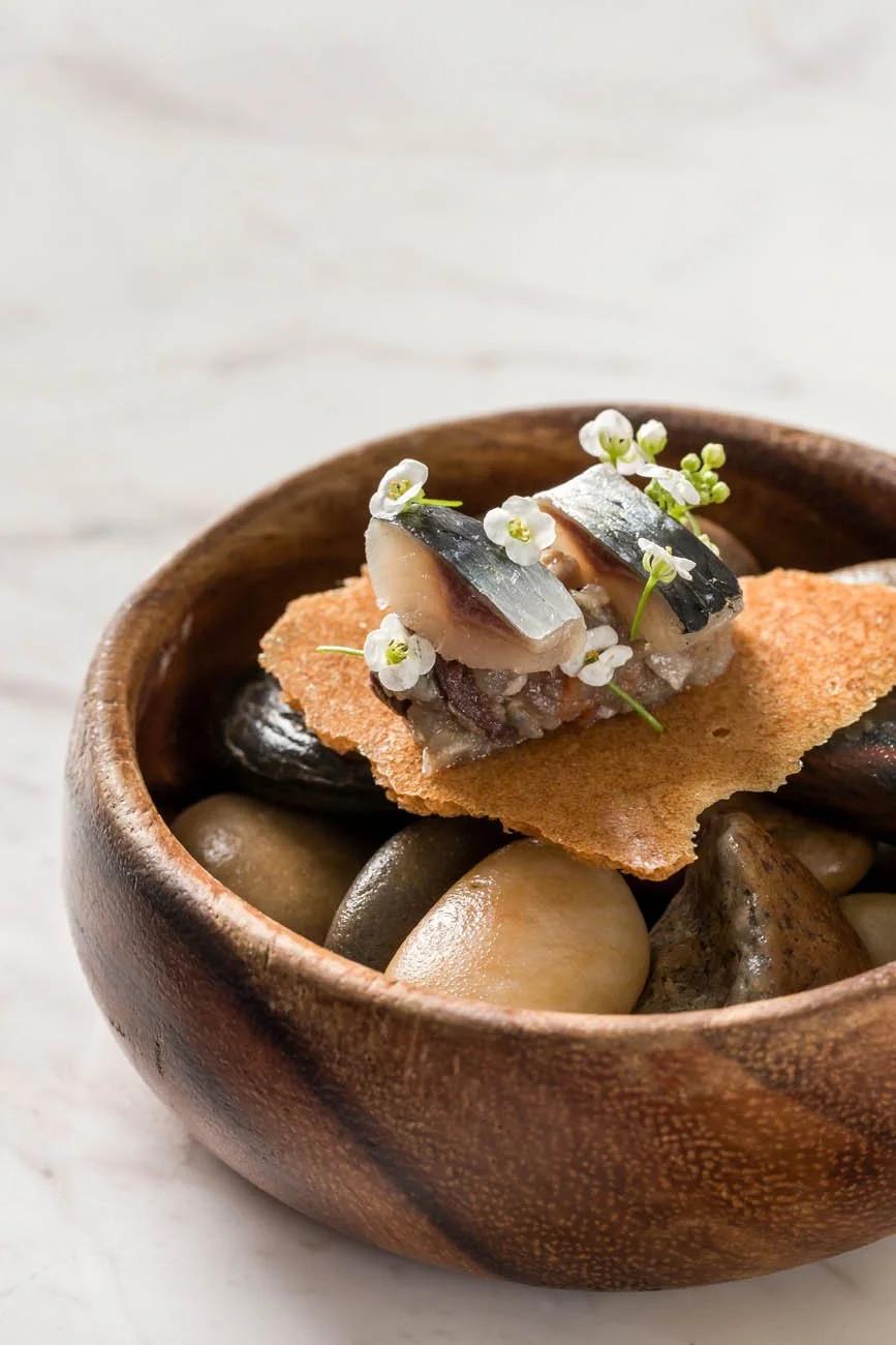 Tigela de madeira com pedras ao fundo, contendo peixe, uma camada fina e crocante, adornada com flores brancas pequenas.