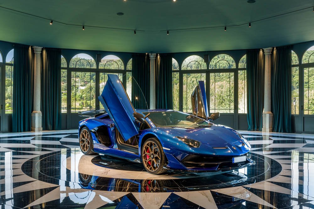 Carro esportivo sedã de cor azul com portas em tesoura abertas exibido em um salão com piso preto e branco e janelas grandes ao fundo.