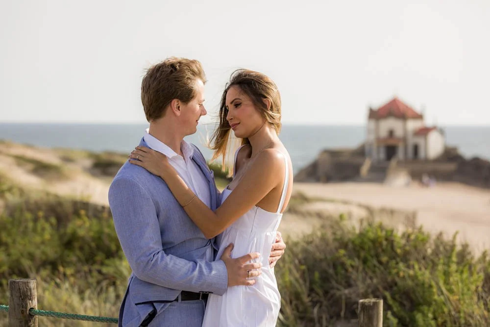 Um casal se abraçando na praia, com uma casa ao fundo na praia.