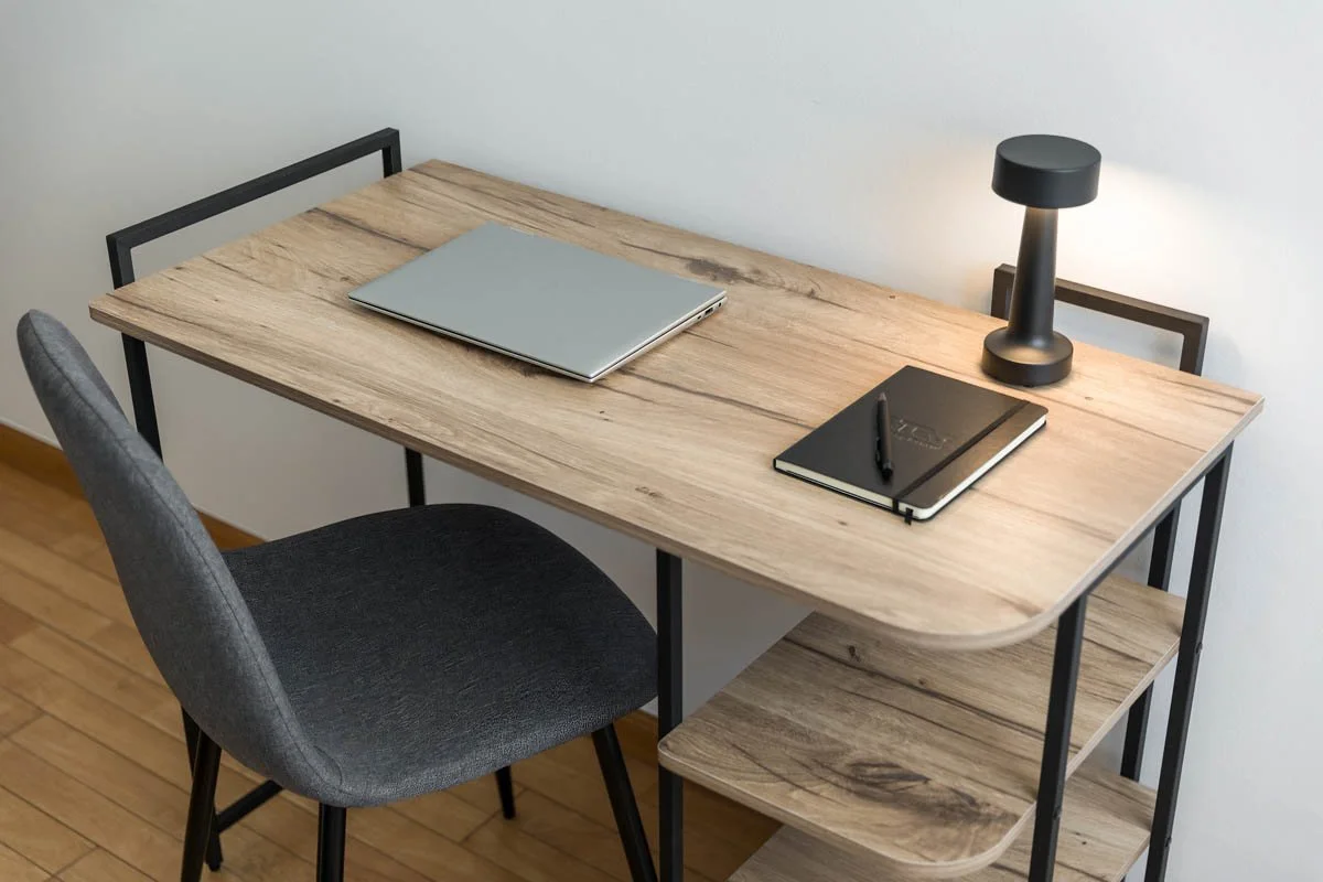 Mesa de estudo de madeira clara com iluminação de mesa, laptop, caderno e caneta, ao lado de uma cadeira cinza