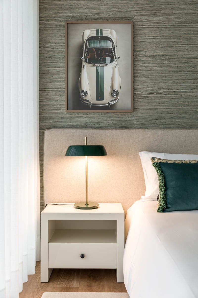 Quarto com cabeceira bege, luminária verde escura, quadro com foto de carro clássico vermelho e branco com faixa verde,; cama com lençol branco, travesseiros verdes e decorativos.