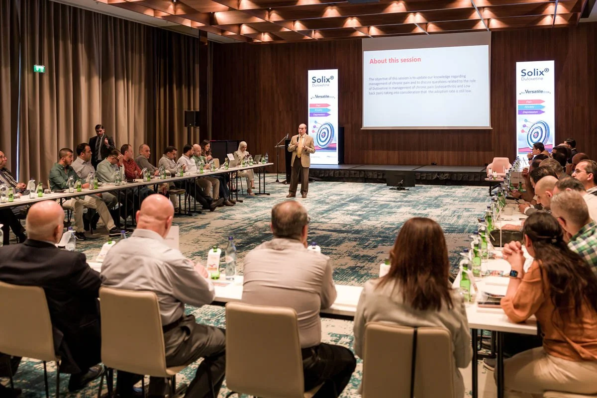 Reunião ou conferência empresarial com várias pessoas sentadas ao redor de uma mesa em formato de U, ouvindo uma apresentação de um homem de terno na frente de uma tela de projeção que exibe informações sobre uma sessão de treinamento ou palestra. As
