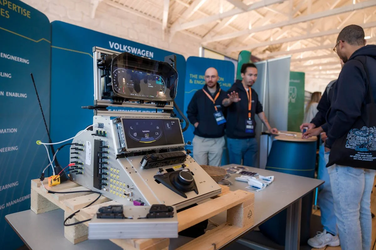 Exposição de tecnologia automotiva com painel digital e simuladores, com pessoas discutindo no estande da Volkswagen em evento técnico