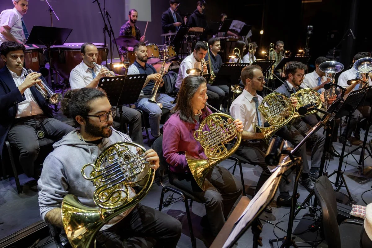 Orquestra de músicos tocando instrumentos de metal e de percussão em um palco.
