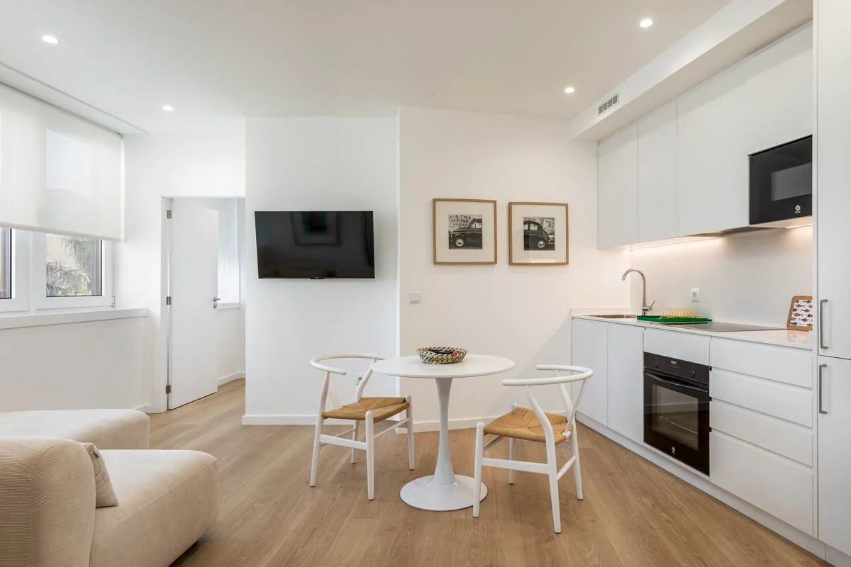 Sala de estar com cozinha integrada, sofá bege, mesa de jantar branca com duas cadeiras de vime, televisão na parede, quadros decorativos, janelas com persianas brancas, piso de madeira clara.