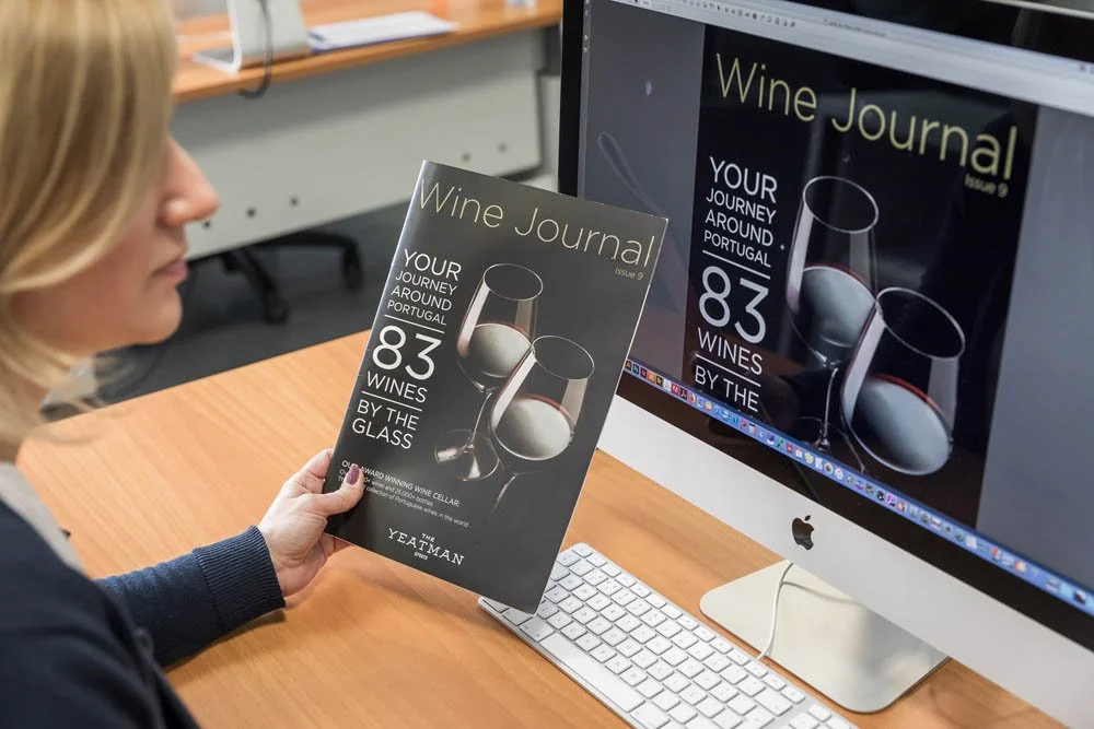Pessoa olhando para uma revista de jornal de vinhos ao lado de um computador Apple. No monitor, uma prévia do jornal com a mesma capa. A capa da revista apresenta uma imagem de duas taças de vinho com uma garrafa ao fundo.