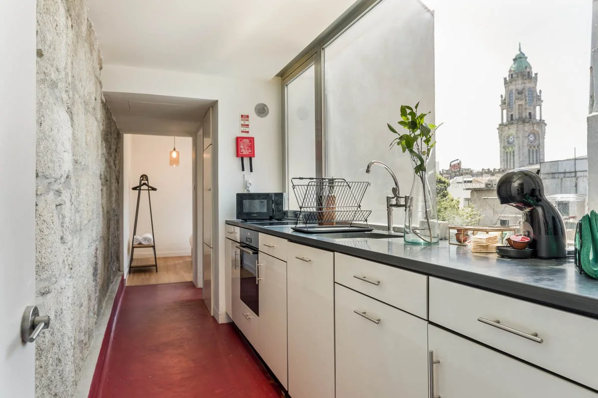 Cozinha com bancada preta, armários brancos, janela grande com vista para uma torre de igreja histórica, vaso com planta, máquina de café e utensílios na bancada, parede de pedra exposta, piso vermelho, iluminação natural.