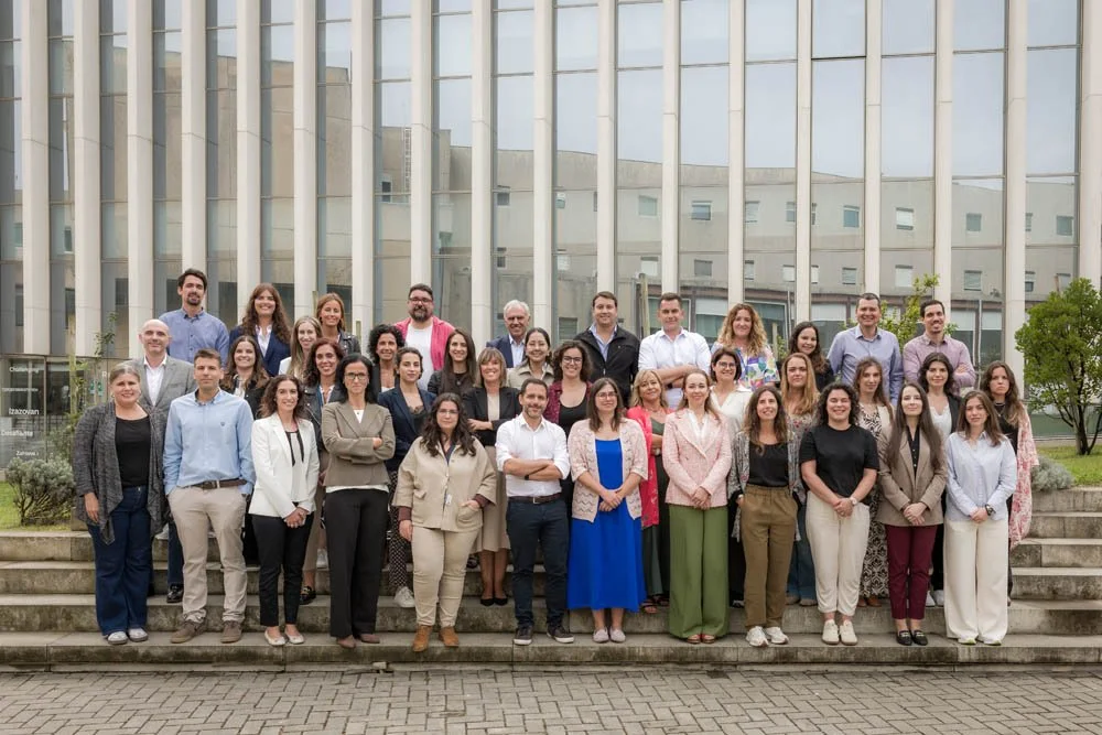 Grupo de pessoas posando em frente a prédio de vidro, sorrindo, em ambiente ao ar livre.