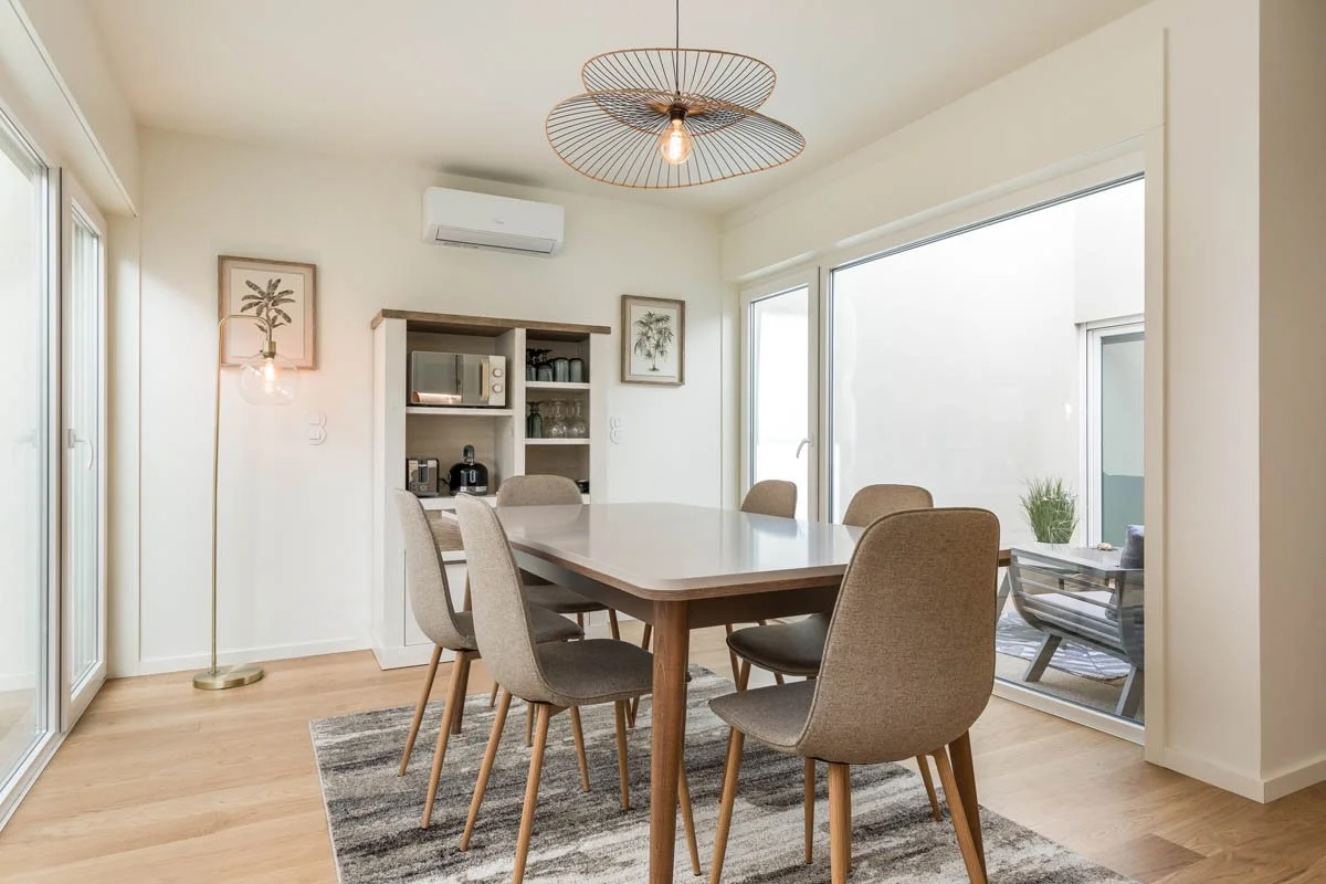 Sala de jantar com mesa de madeira e várias cadeiras confortáveis, janelas grandes com luz natural, decoração minimalista, parede branca, iluminação decorativa moderna.