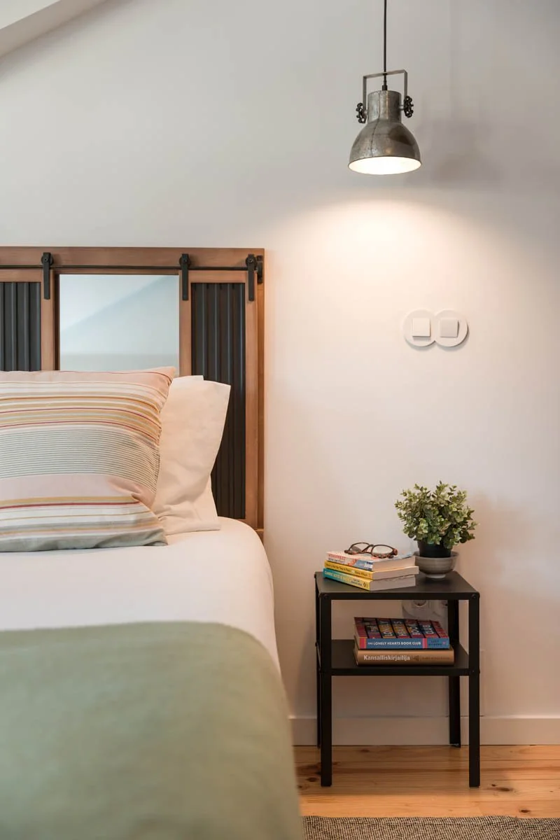 Quarto com cabeceira de madeira, travesseiros, luminária de teto metálica, parede branca, um criado-mudo com livros, óculos, uma planta e uma parede com interruptores.