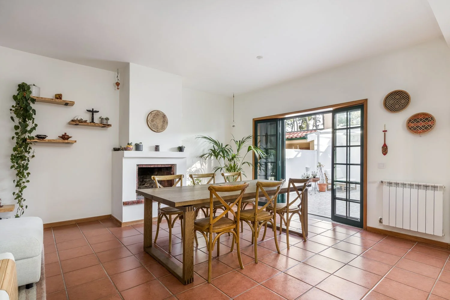 Sala de jantar com mesa de madeira e cadeiras de vime, parede branca com prateleiras de madeira, plantas, decoração tradicional e janela de portas de vidro para o quintal ao ar livre.